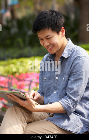 Young Chinese Man Using Digital Tablet Whilst  Relaxing On Park Bench Stock Photo