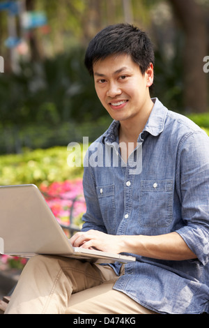 Young Chinese Man Using Laptop Whilst  Relaxing On Park Bench Stock Photo