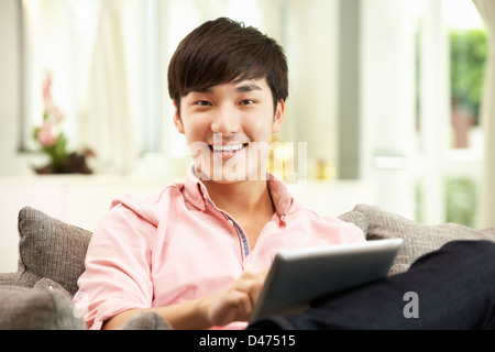 Young Chinese Man Using Digital Tablet Whilst Relaxing On Sofa At Home Stock Photo