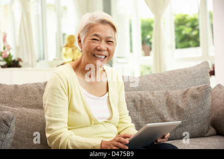 Senior Chinese Woman With Tablet Computer Whilst Relaxing On Sofa At Home Stock Photo