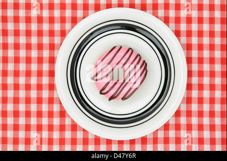 A donut with pink icing and drizzled chocolate, on a white plate which is placed on a red-and-white checkered tablecloth, studio shot taken, top view. Stock Photo