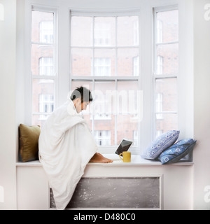 Japanese young man using laptop while sitting on window sill and drinking coffee on a cold day in London Stock Photo