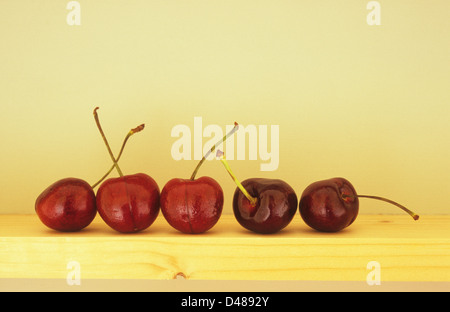 Eye-level view of five dark red cherries with stalks lined up on edge of narrow wooden shelf Stock Photo