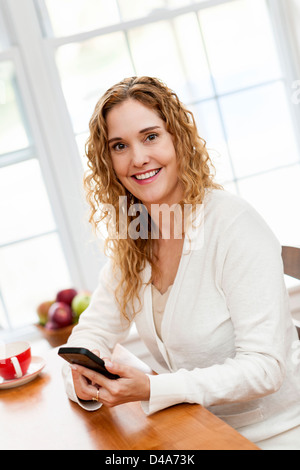 Portrait of smiling woman holding cell phone sitting at table in home kitchen by window Stock Photo