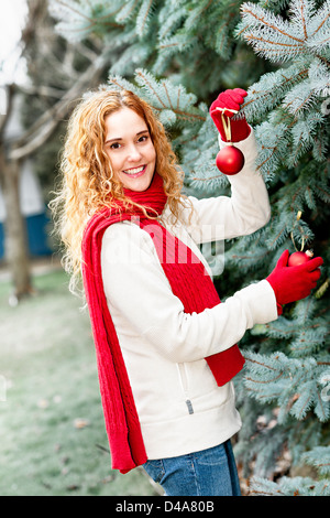 Joyful woman hanging Christmas ornaments on spruce tree outdoors in yard near home Stock Photo