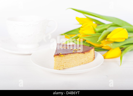 piece of layer cake with chocolate on a white plate, in the background a cup of tea and a bouquet of yellow tulips Stock Photo