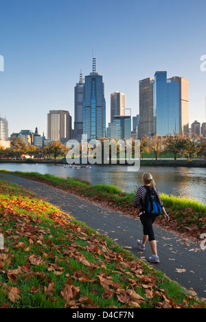 Woman walking on banks of Yarra River with city skyline in background. Melbourne, Victoria, Australia Stock Photo