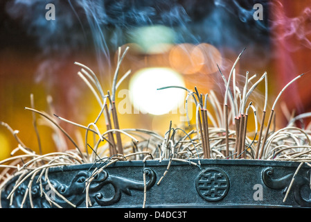 Close-up view of burning incense sticks in bowl with smoke rising over blurry background. Traditional rituals at Buddhist temple Stock Photo