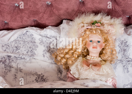 Face of a beautiful vintage doll with her long blonde hair in ringlets and wearing pearls and a fancy hat lying on a bed Stock Photo