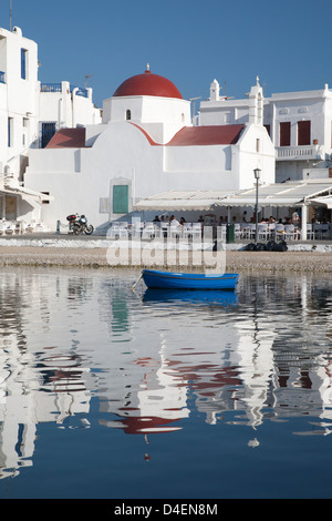 White church with red dome, outdoor cafés, boat and reflections in the water of Mykonos harbour, Greek Islands Stock Photo