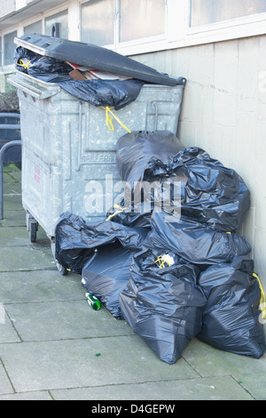 Rubbish overflowing from a bin. The waste is in tied black bin bags, some are in a galvanised wheelie bin, others are piled on the ground. Stock Photo