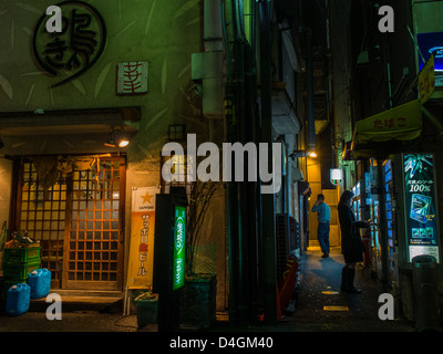 Osaka at night.  In an alley next to a restaurant a woman buys cigarettes from a vending machine while a man talks on his phone. Stock Photo