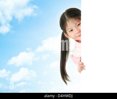 Young Asian girl hiding behind a blank white card in summer day, blue sky as background. Stock Photo