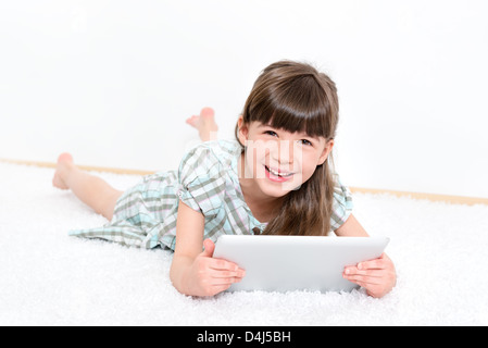 Joyful pretty little girl laughing as she lies on her stomach on a white carpet playing with a modern digital tablet  in a room.