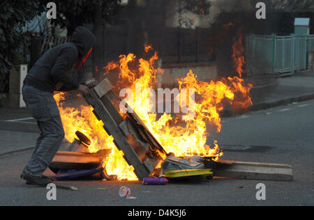A hooded anti-NATO activist throws a wooden pallet into a fire at a ...