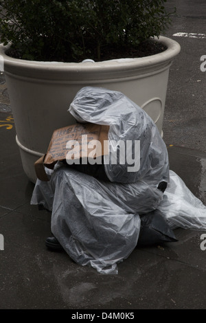 Homeless man wrapped in plastic simultaneously begging and fighting off the rain, Lexington Ave., Manhattan. Stock Photo