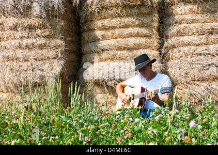 A cowboy sits in front of a wall of hay bales playing a guitar with summer field flowers in the foreground Stock Photo