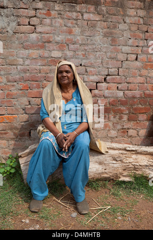 A traditionally dressed Punjabi woman in an embroidered salwar kameez sitting on a log outside the family home in North India Stock Photo