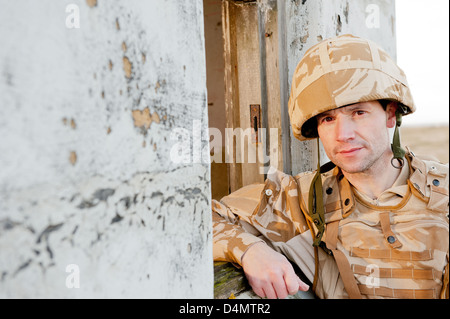 British soldier wearing desert camouflage, looking at the camera and leaning against a derelict building. Stock Photo