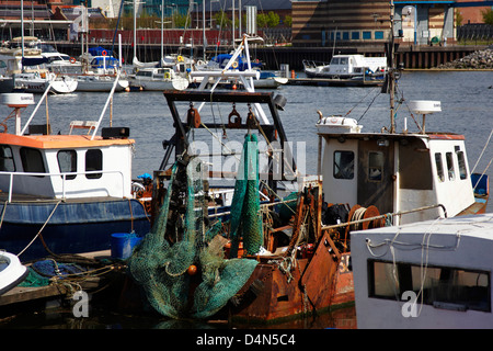 Small old fishing boat with net and hoists moored in modern marina with yachts in the background Stock Photo