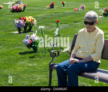 An elderly woman wearing sunglasses sitting alone on a bench in a cemetery looking sad. Stock Photo
