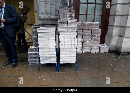 Stacked newspapers on a newsstand outside underground tube station and businessman in suit reading paper Central London England UK  KATHY DEWITT Stock Photo
