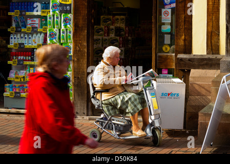 Elderly woman in mobility scooter reading a newspaper outside a shop in Stafford England UK Stock Photo