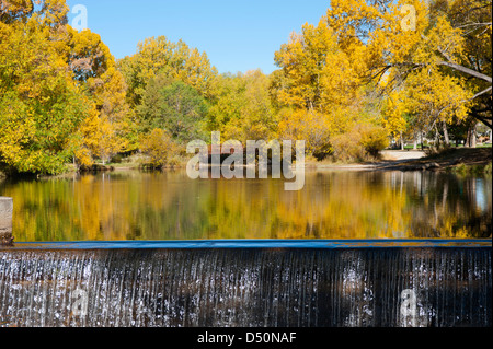 A small lake, complete with footbridge, reflects the harvest colors of Autumn. The waterfall in the foreground adds symmetry. Stock Photo