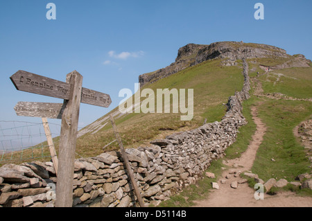 Timber fingerpost or waymark sign by stone wall, pointing to footpath route up to rocky crag summit of Pen-y-ghent -Yorkshire Three Peaks, England, UK Stock Photo