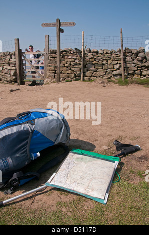 By summit of Pen-y-ghent, close-up of rucksack, ordnance survey map case & trekking pole on track (walkers by fingerpost beyond) - Yorkshire Dales, UK Stock Photo