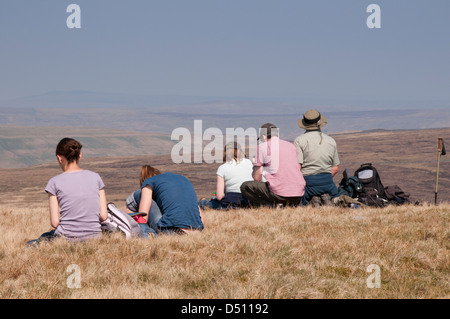 Rear view of people (walkers) sitting on high summit of Pen-y-ghent (one of Yorkshire Three Peaks) looking out over scenic hazy uplands - England, UK. Stock Photo