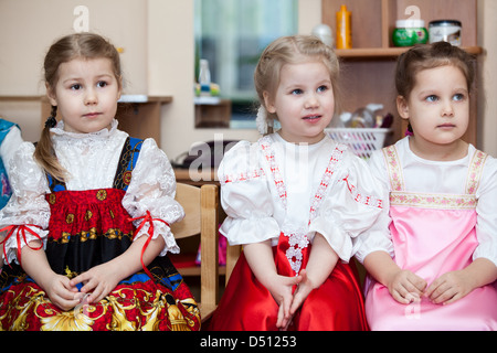 Cute girls in Russian traditional clothes in kindergarten sitting on the chairs in classroom. Russia Stock Photo