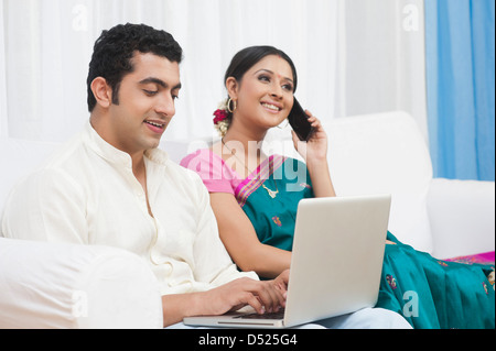 Man using a laptop with his wife talking on the mobile phone beiside him Stock Photo