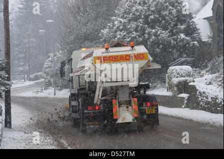 It is snowing heavily during spell of cold, snowy winter weather & council gritter wagon (with snow plough) is driving along, clearing main road through village, spreading rock salt on slippy surface - Hawksworth, West Yorkshire, England, UK. Stock Photo