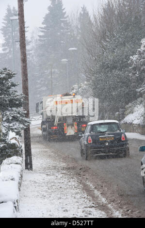 It is snowing heavily during spell of cold, snowy winter weather & council gritter wagon (with snow plough) is driving along, clearing main road through village, spreading rock salt on slippy surface - Hawksworth, West Yorkshire, England, UK. Stock Photo
