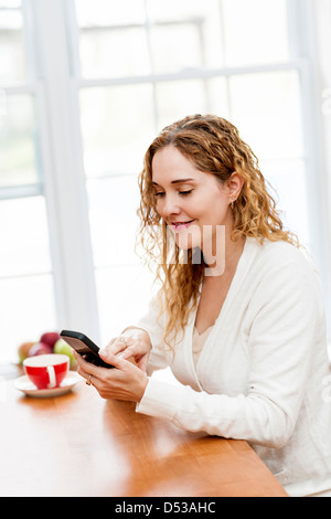 Portrait of smiling woman holding cell phone sitting at table in home kitchen by window Stock Photo
