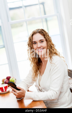 Portrait of smiling woman holding cell phone sitting at table in home kitchen by window Stock Photo