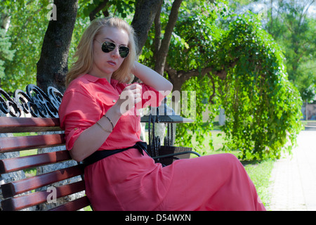 the beautiful girl in a red dress sits on a bench in a summer garden Stock Photo