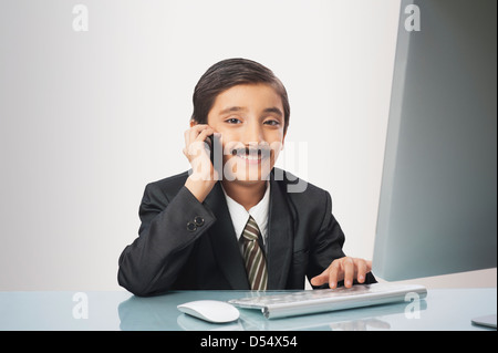 Boy imitating like businessman talking on a mobile phone while working on a desktop pc Stock Photo