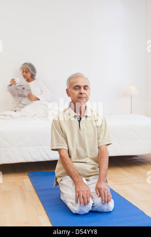 Senior man practicing yoga with his wife reading newspaper on the bed Stock Photo