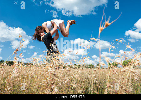 Happy young couple standing in open field of long grass with the young man lifting the gorgeous girl into the air, kissing. Stock Photo