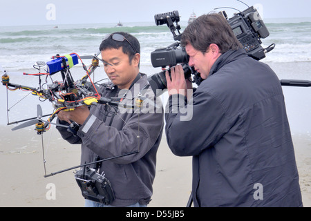 A technology demonstration on a beach in 2013 featuring a man holding a multi-rotor camera drone while a cameraman films the device. Stock Photo
