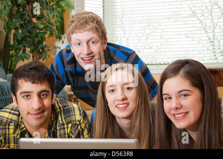 A diverse group of teenagers gathered around a laptop in a home setting looking at the camera Stock Photo