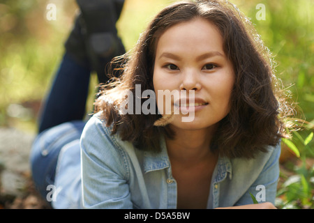 Beautiful young Asian model wearing denim shirt laying on ground in lush foliage on sunny day. Stock Photo