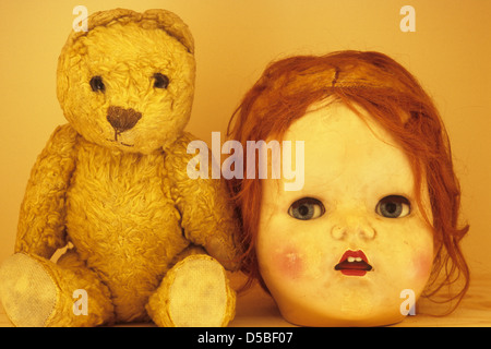 Close up of shelf with battered old teddy bear next to head of doll with big blue eyes open mouth and untidy ginger hair Stock Photo