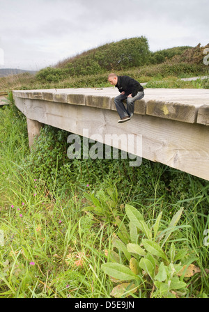 Photomontage of a young man sitting on a wooden walkway in nature and looking down. Man is small compared to the surrounding env Stock Photo