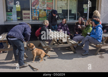 Walking friends enjoy a rest on benches at an outdoor cafe in Epping Forest, Essex, England. Gathered on bench seats and wrapped up against a Spring chill, the people sit with foam cups of tea, talking next to another person whose pet American Staffordshire Terrier is on a lead. The outdoor cafe is in a car park inside Epping Forest, an area of ancient woodland in south-east England, straddling the border between north-east Greater London and Essex. It covers 2,476 hectares and contains areas of woodland, grassland, heath, rivers, bogs and ponds - popular with families and more serious walkers Stock Photo