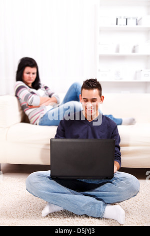 Happy man using laptop on the floor, while his girlfriend is sitting on the sofa bored, sad and upset Stock Photo