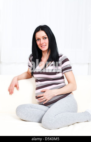 Beautiful young pregnant woman with long black hair smiling and sitting on the sofa Stock Photo