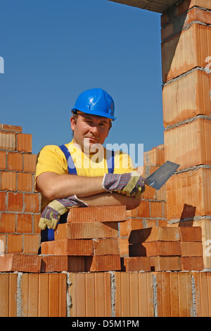 Construction worker in yellow t-shirt and blue helmet holding stainless steel trowel posing among unfinished brick house walls Stock Photo